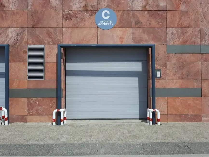 Closed gray sectional loading dock door in a marble-clad wall with red and white safety bollards