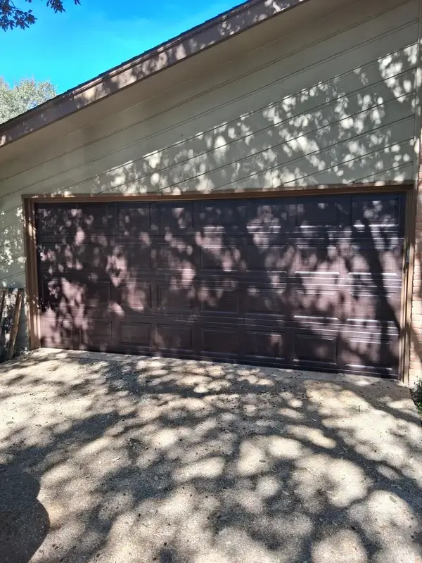 Dark brown raised-panel double garage door on a tan house with tree shadows on the driveway