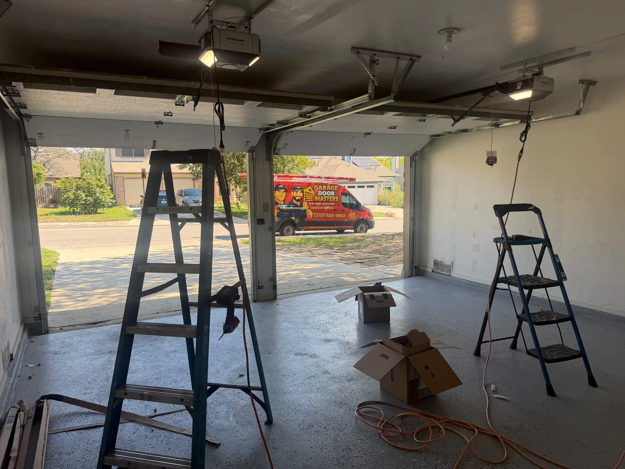 Garage work area with open doors, ladders, equipment boxes, electrical cords, and a service vehicle outside.