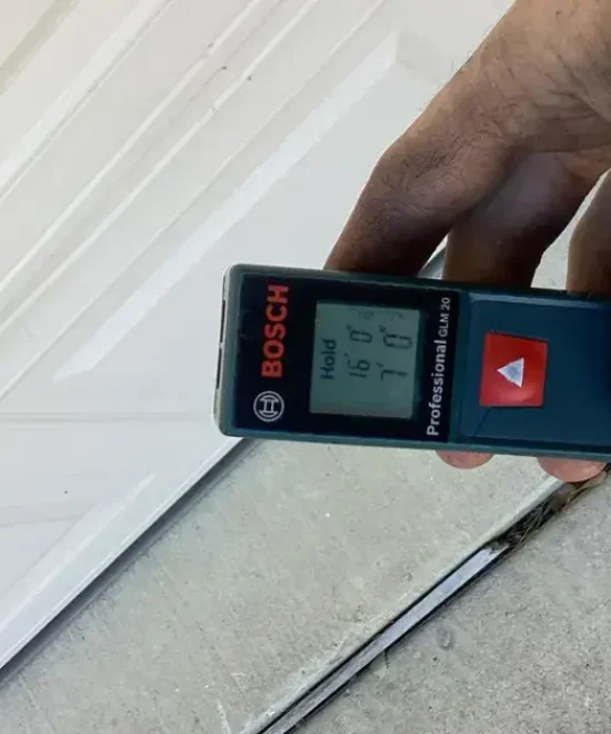 Hand holding a Bosch laser distance meter against the bottom edge of a white raised-panel garage door