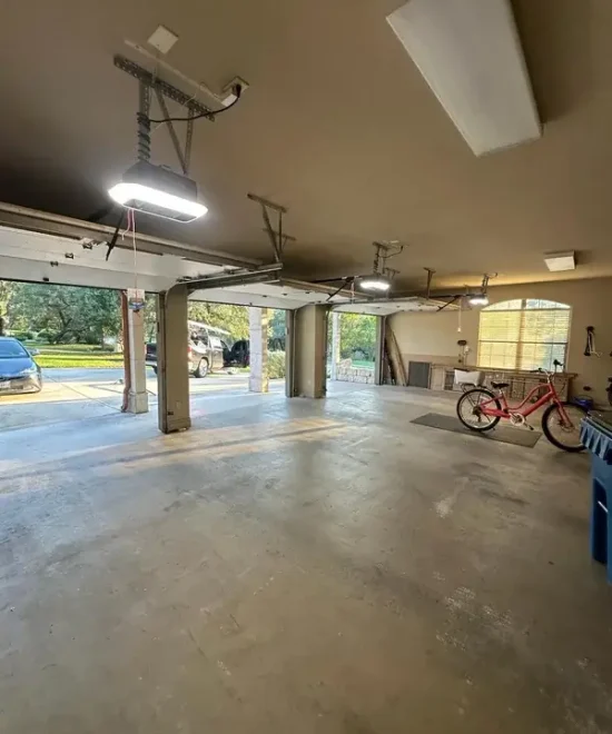 Empty residential garage interior with open doors, ceiling-mounted openers overhead, and a red bicycle by a workbench
