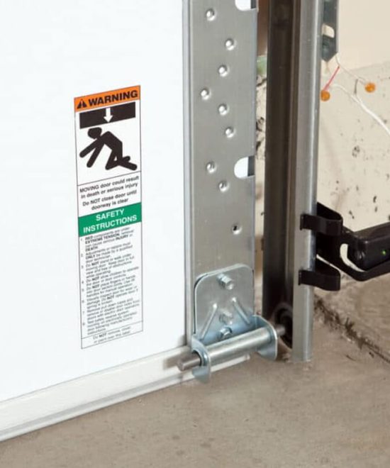 Photo-eye safety sensor with a green light on a garage door track, next to a warning decal on the door panel