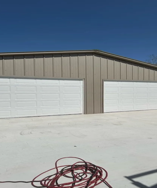 Tan metal shop building with two closed white raised-panel garage doors and a coiled extension cord on the concrete