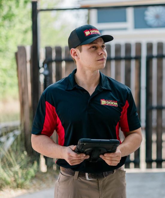 Technician in a Garage Door Masters cap and polo holding a rugged tablet by a wooden fence
