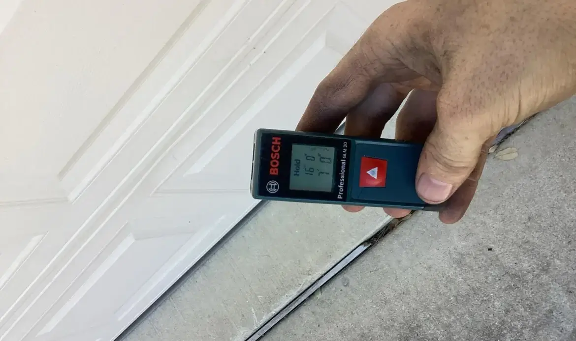 Hand holding a Bosch laser distance meter at the bottom of a white garage door, display reading 16 feet by 7 feet