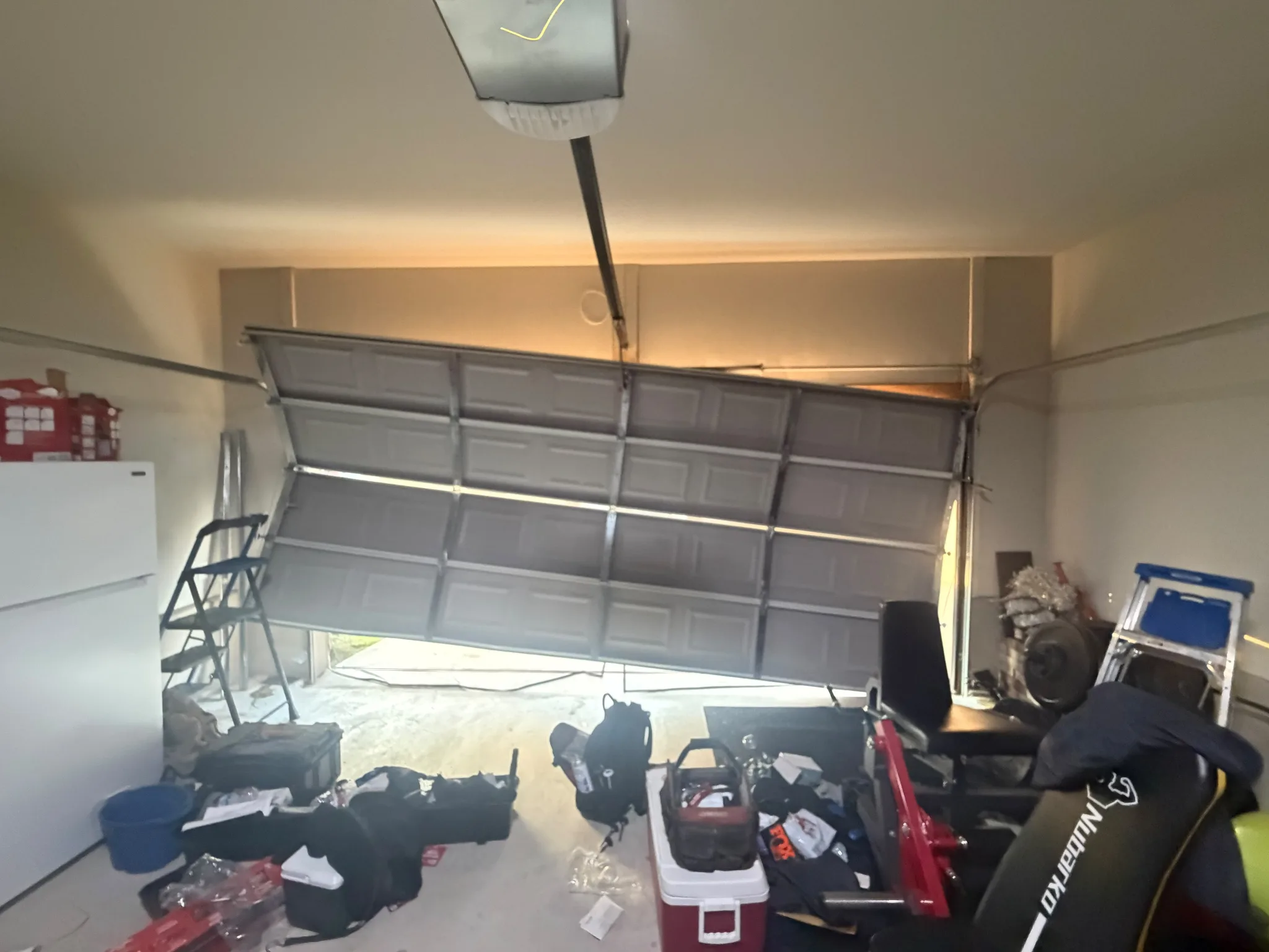 Partially open gray sectional garage door beneath a ceiling-mounted opener, with tools and equipment on the floor.
