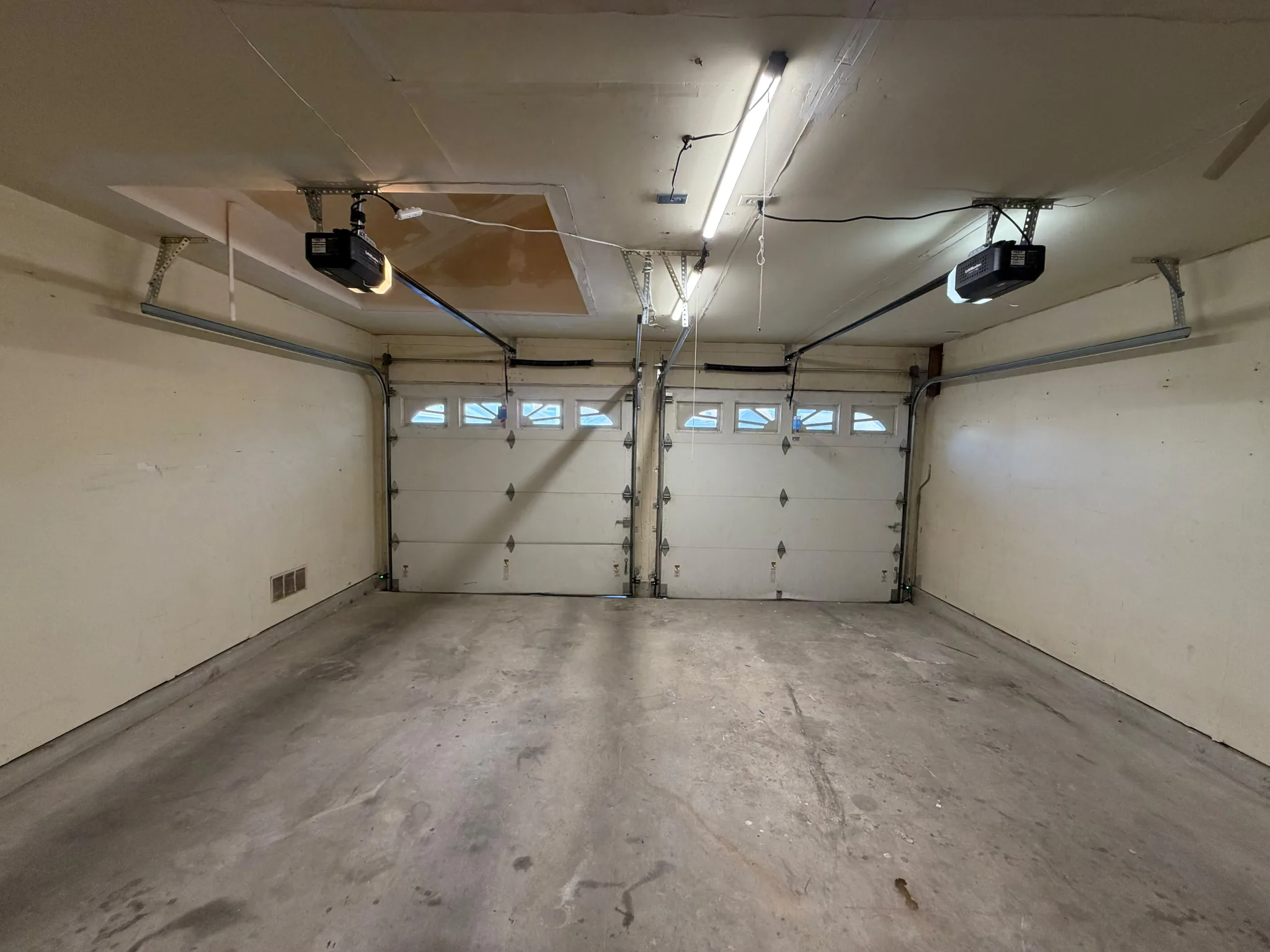 Two white sectional garage doors with small windows, overhead tracks, and ceiling-mounted openers in an empty garage.