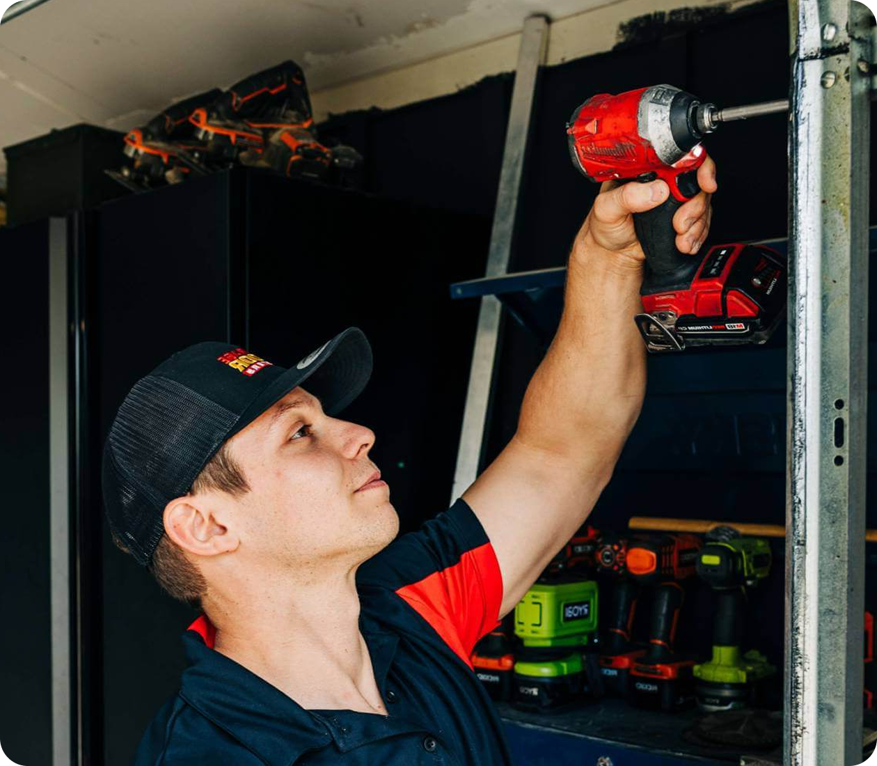Technician in a branded cap raising a red cordless impact driver toward a galvanized steel door track