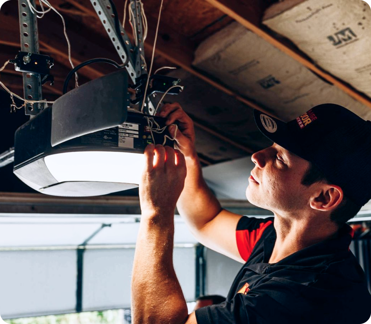 Technician in a black cap reaching up to wire the terminals of a ceiling-mounted garage door opener