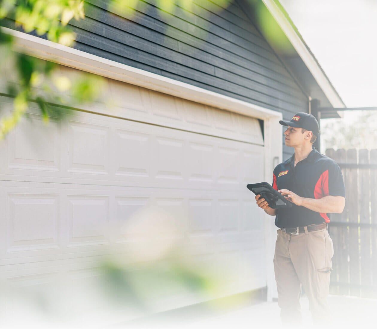 Technician in a black and red uniform holds a tablet beside a white raised-panel garage door