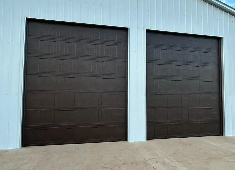 Two tall brown woodgrain garage doors, both closed, on a white metal shop building with a concrete apron