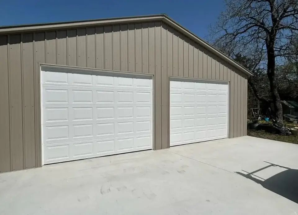 Two white raised-panel garage doors on a tan metal workshop building under a blue sky