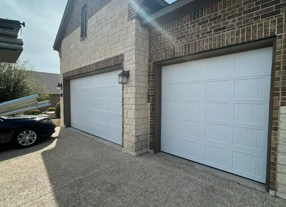 Two closed white raised-panel garage doors on a brick and stone home with a carriage lamp and aggregate driveway